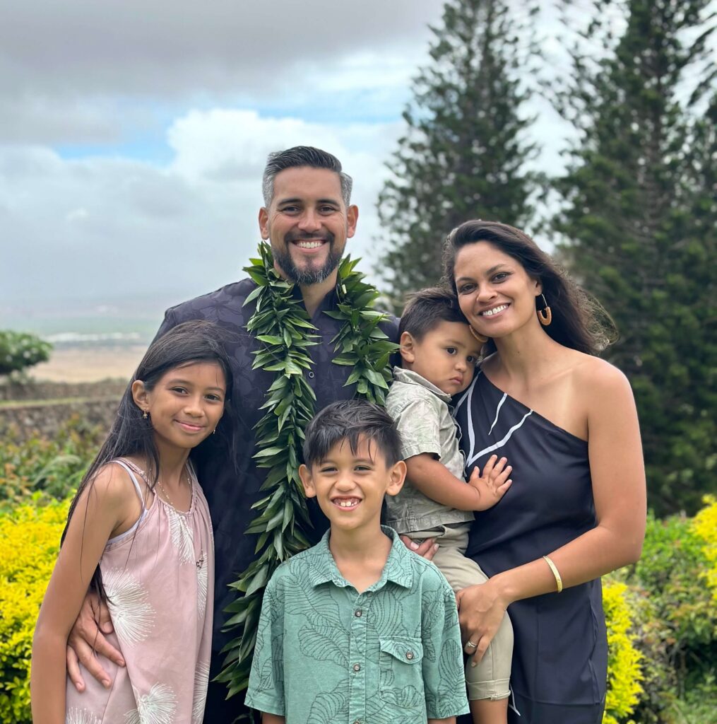 Family of five posing outdoors, wearing leafy leis, with trees and yellow flowers in the background.